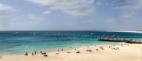 A beach with clear turquoise water, a pier with people, and boats.