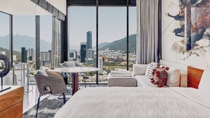 Habitació d'hotel moderna amb un llit gran, zona d'estar amb taula i vista a la ciutat a través de grans finestres.