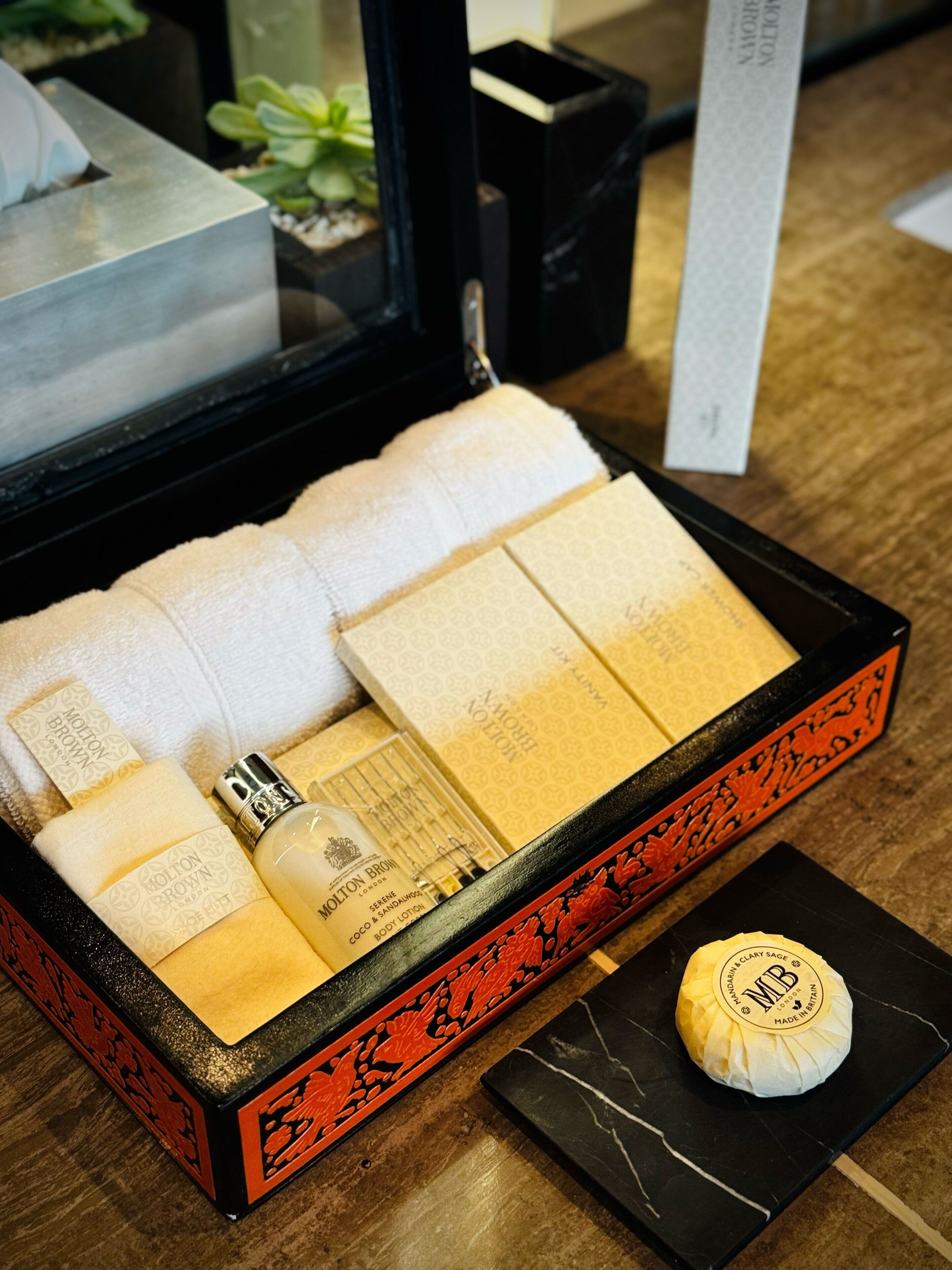 A luxury toiletry set in a black box with a red floral design, including a bottle of body lotion, a bottle of perfume, and several folded towels.