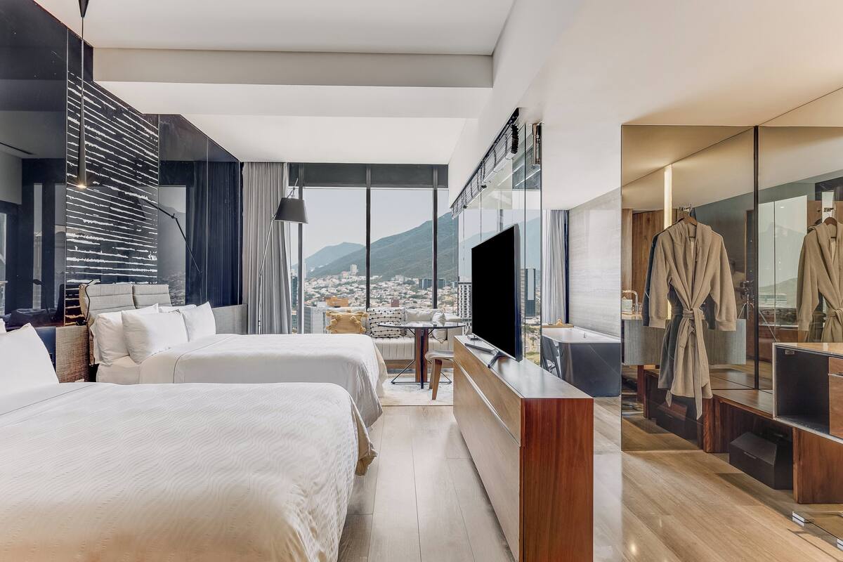 Habitación de hotel moderna con dos camas, televisión, tocador con espejo y vistas a la ciudad y las montañas.