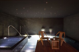 A modern spa room with a hot tub, two wooden chairs, and a small table with a plant.