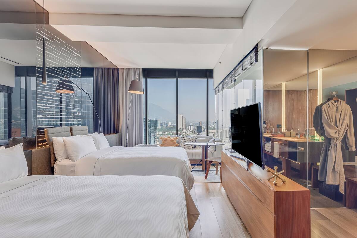 Una habitación de hotel moderna con dos camas, televisión, baño con mampara de vidrio y vistas a la ciudad.