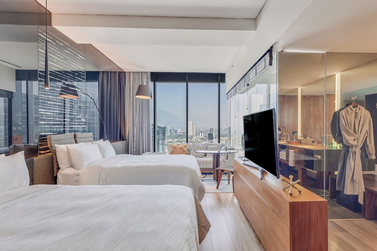 A modern hotel room with two beds, a TV, a glass-enclosed bathroom, and a city view.