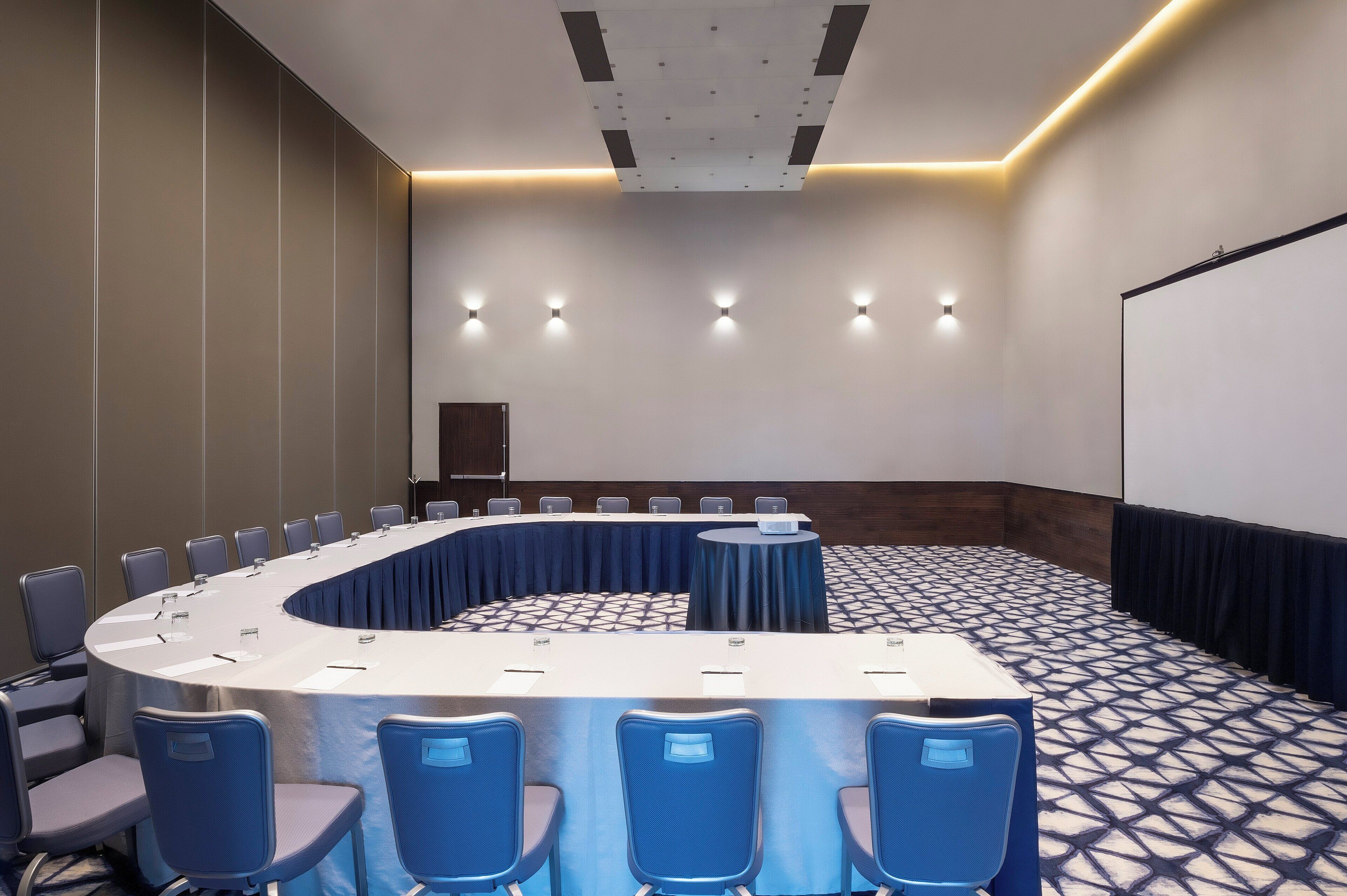 Un salón de conferencias con una mesa curva, sillas azules y una pantalla de proyector.