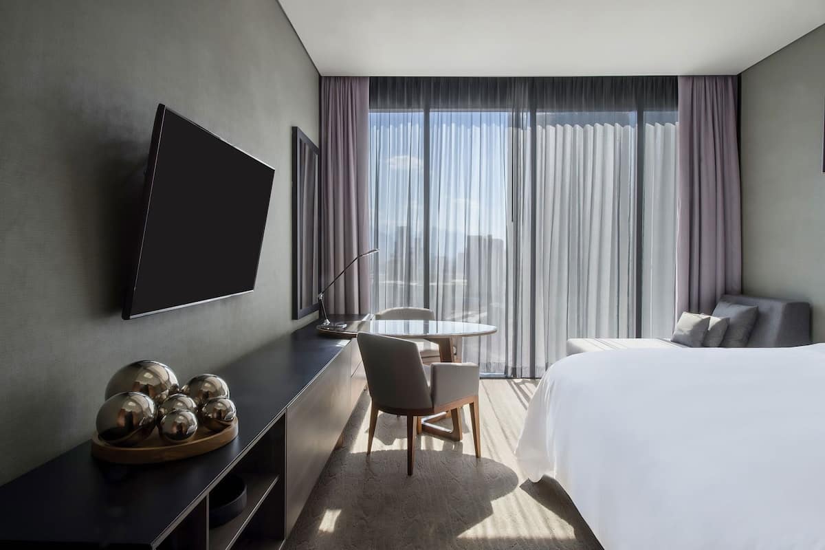Habitación de hotel con cama, escritorio con televisor de pantalla plana, una silla, una mesita y vistas de la ciudad a través de ventanas grandes.