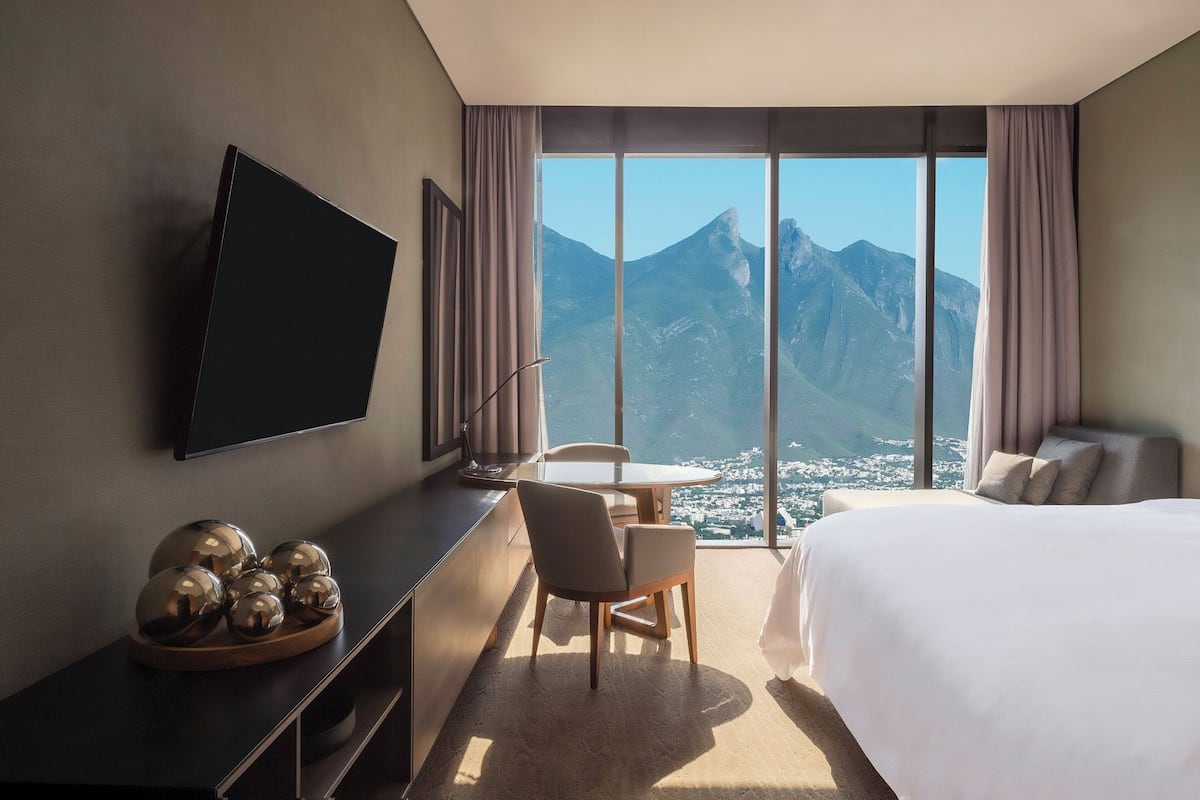 Habitación de hotel con cama, escritorio, silla, televisor y ventana grande con vista a la montaña.