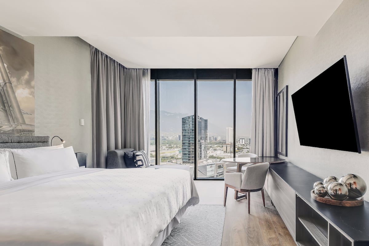 Habitación de hotel moderna con una cama grande, un escritorio, una silla, un televisor de pantalla plana y vistas a la ciudad.