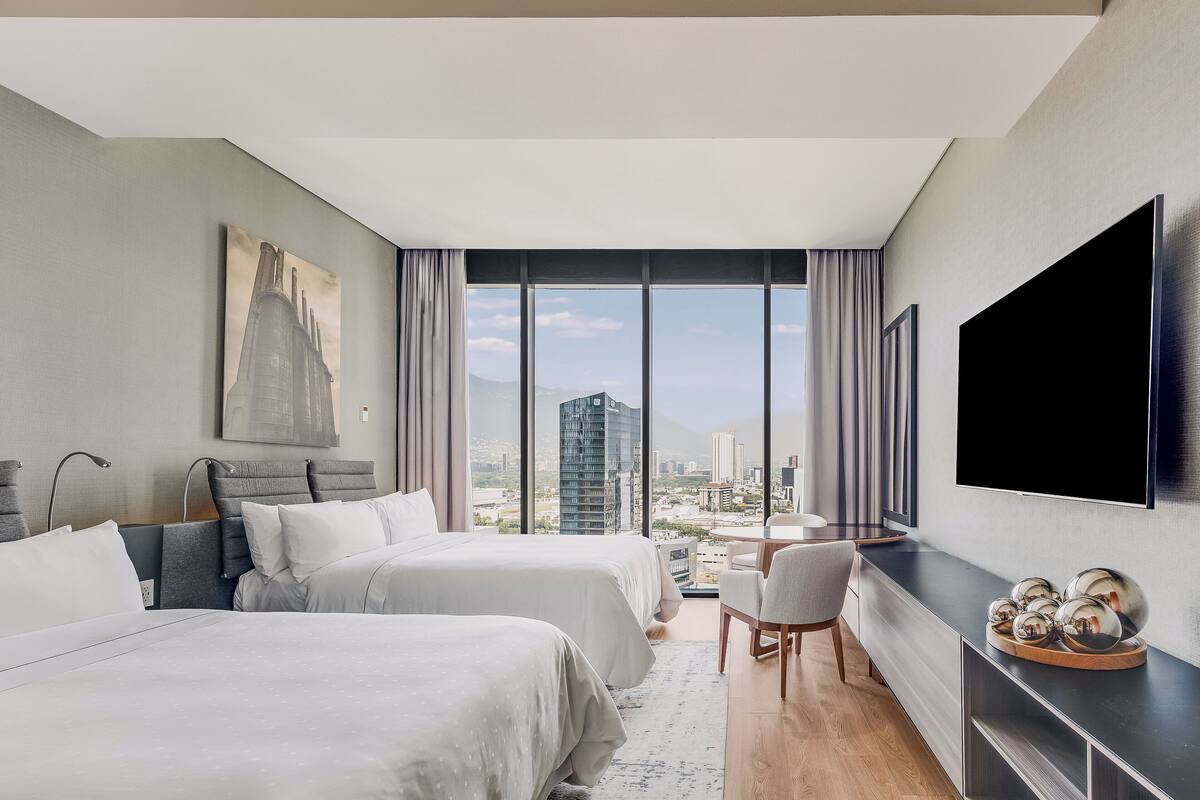 Una habitación de hotel moderna con dos camas, un ventanal con vista a la ciudad, un televisor de pantalla plana y una mesita de noche de madera.