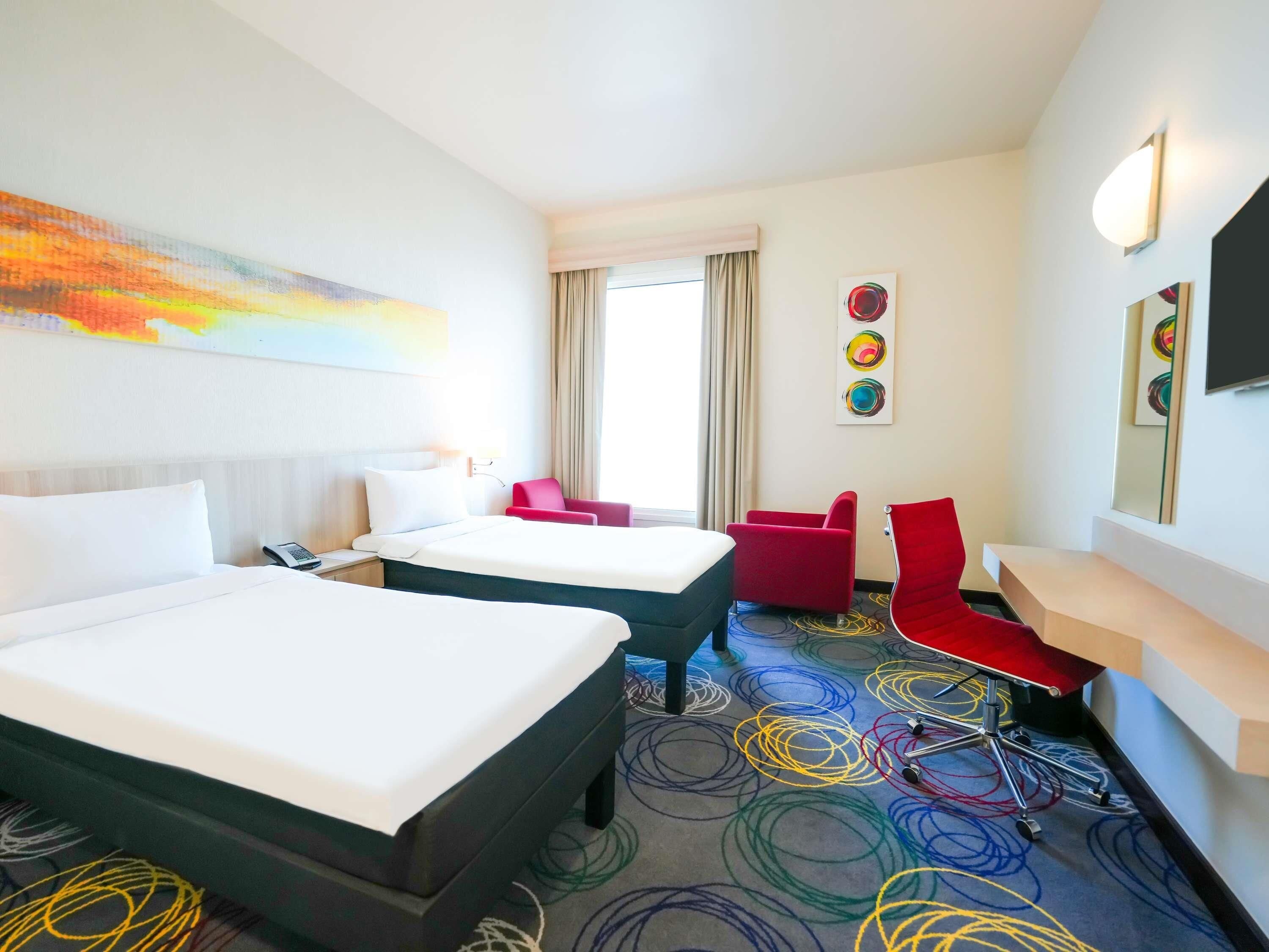 A hotel room with two beds, a desk, a chair, and a large window.