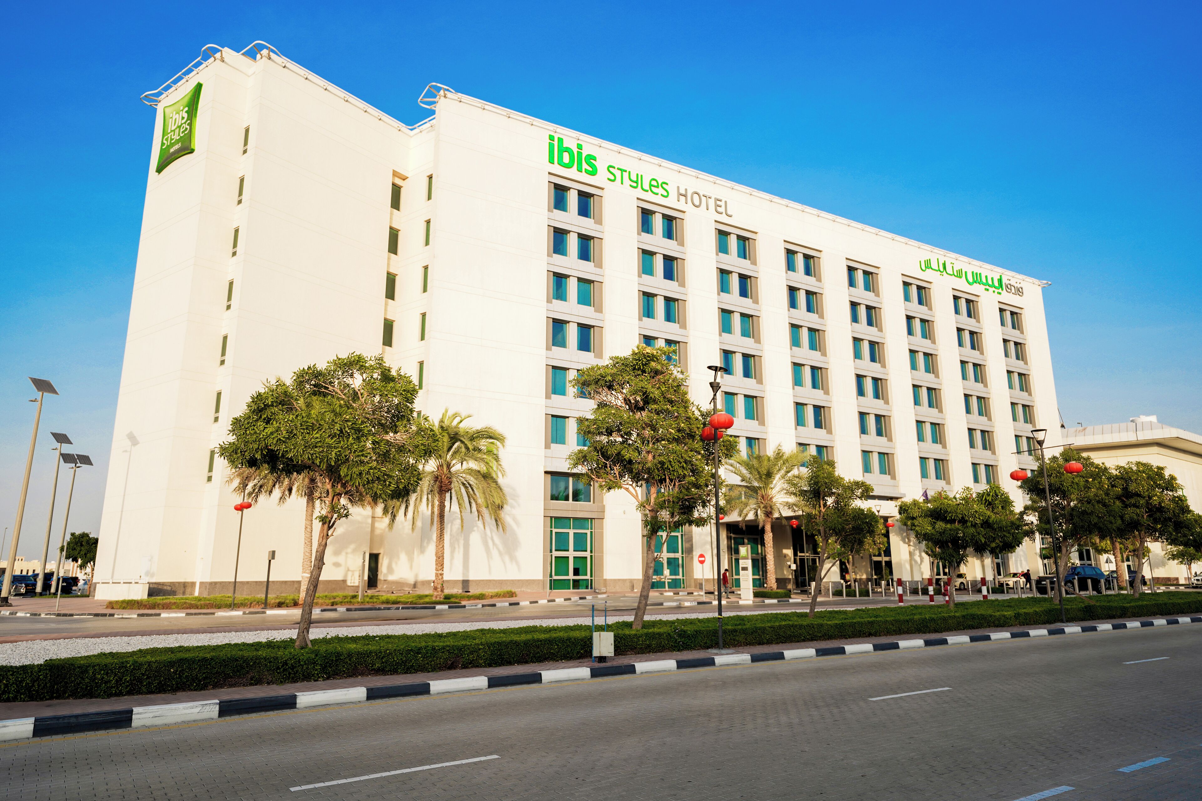 A modern hotel with a prominent "ibis Styles Hotel" sign, featuring a clean facade, multiple floors, and a well-maintained landscaped area with palm trees.