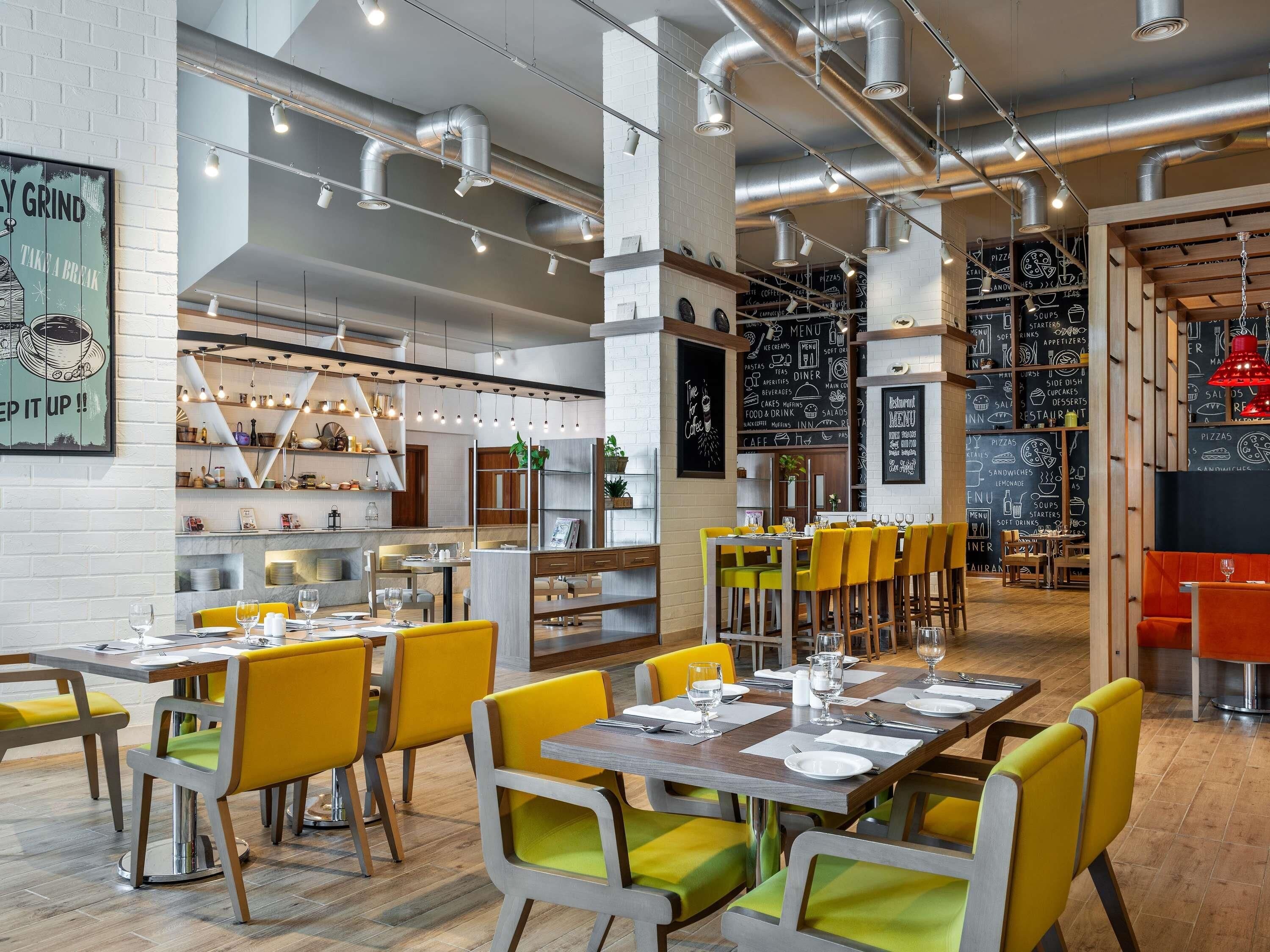 Interior de restaurante com tetos altos, tubulações aparentes e uma mistura de cadeiras amarelas e verdes.