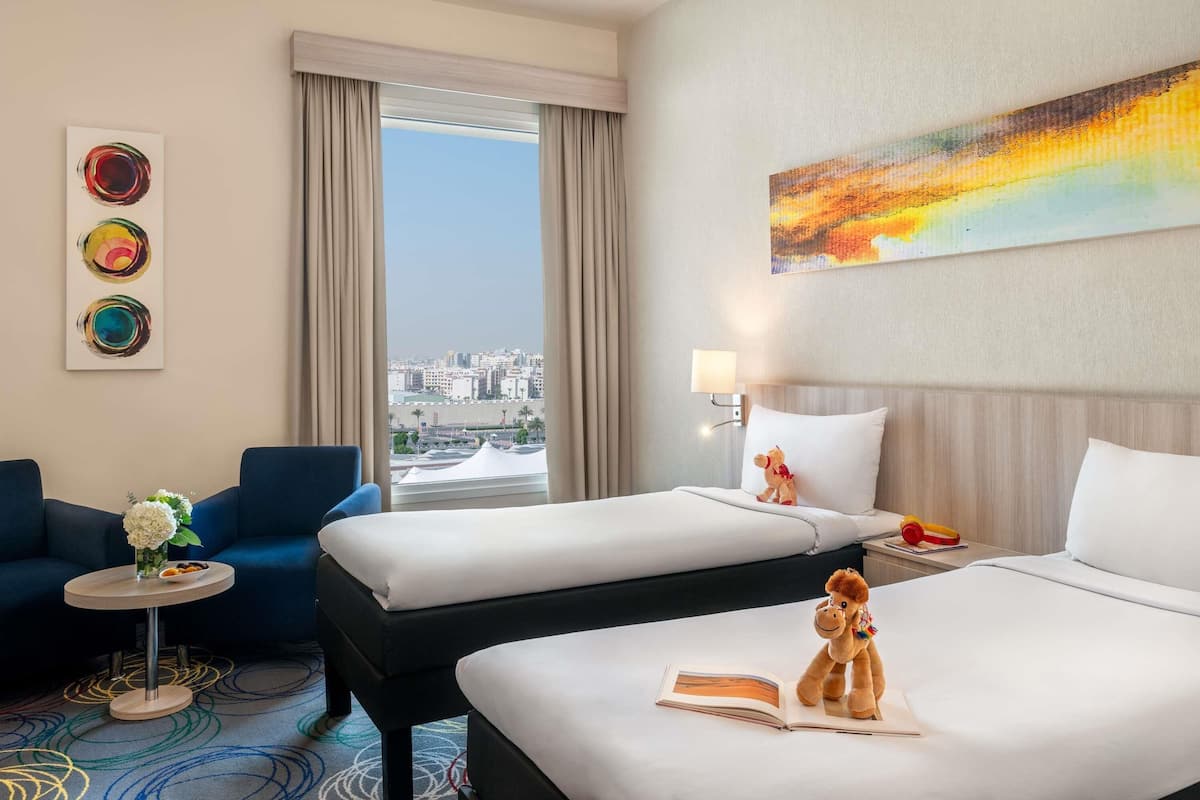 Une chambre d’hôtel avec deux lits, un canapé, une petite table ornée d’un vase de fleurs et une vue sur la ville.