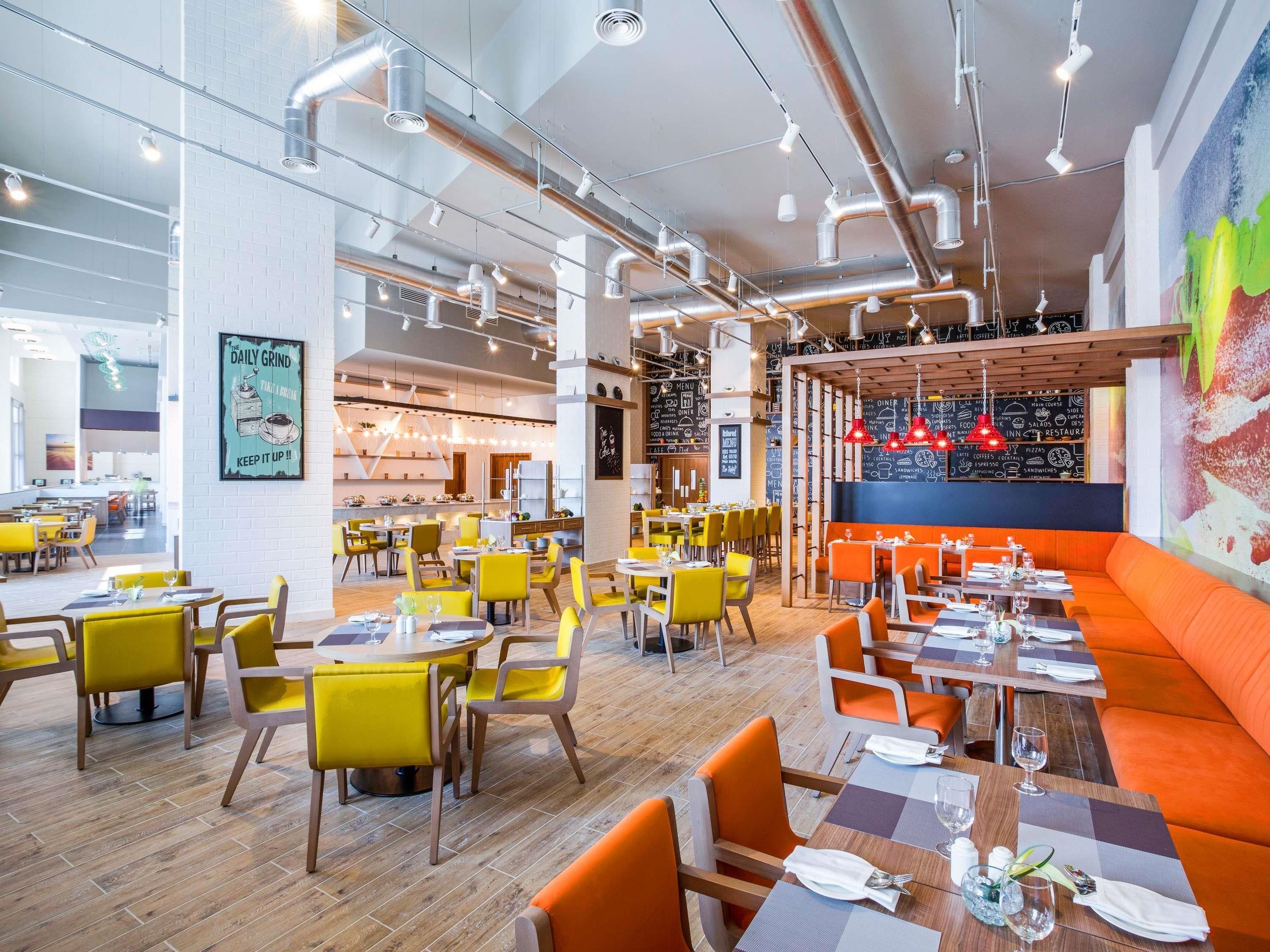 A modern restaurant interior with high ceilings, exposed ductwork, and a mix of yellow and orange seating areas.