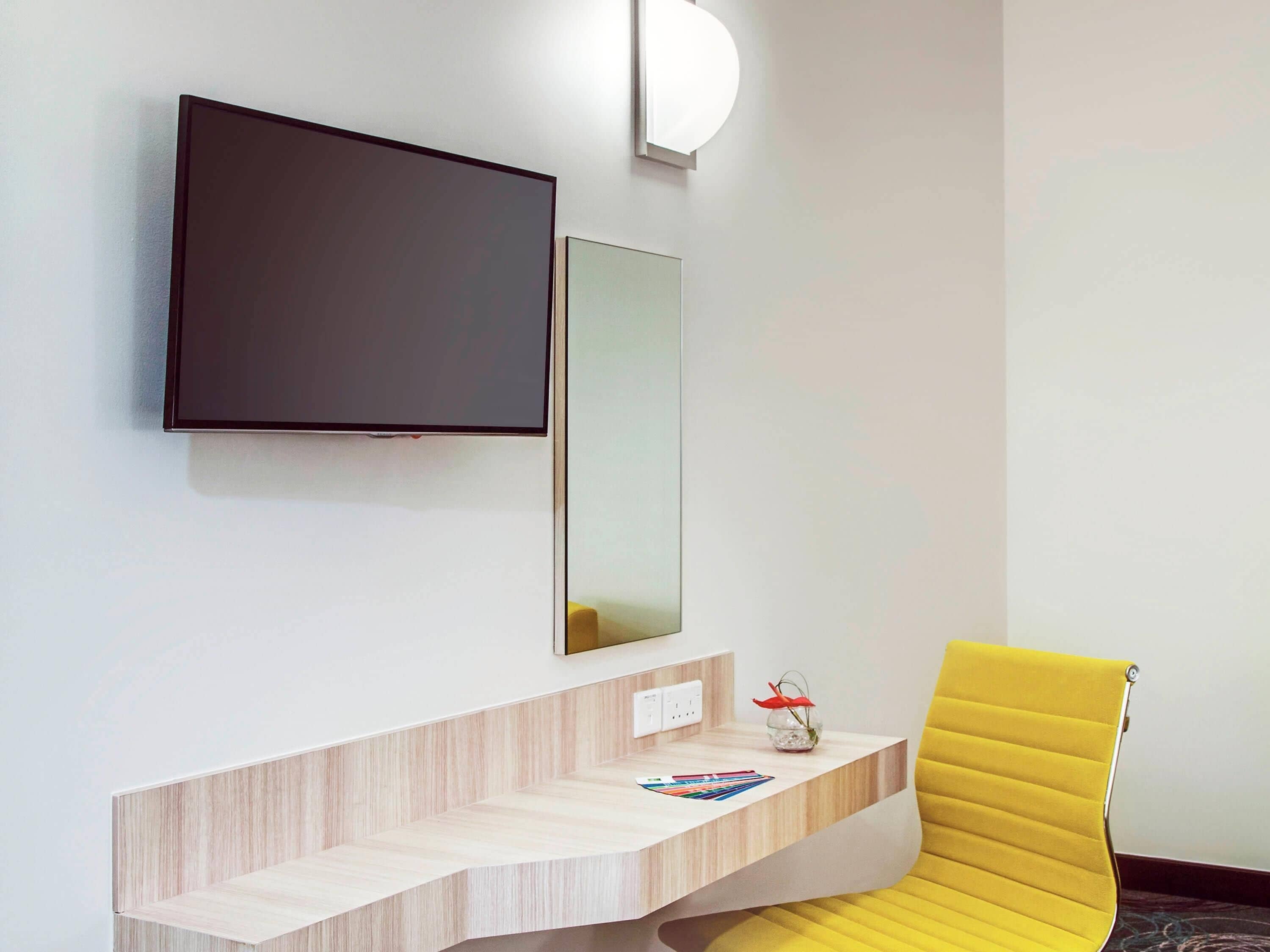 Una habitación de hotel moderna con una televisión de pantalla plana montada en la pared, una mesita de noche con un florero y un sillón amarillo.