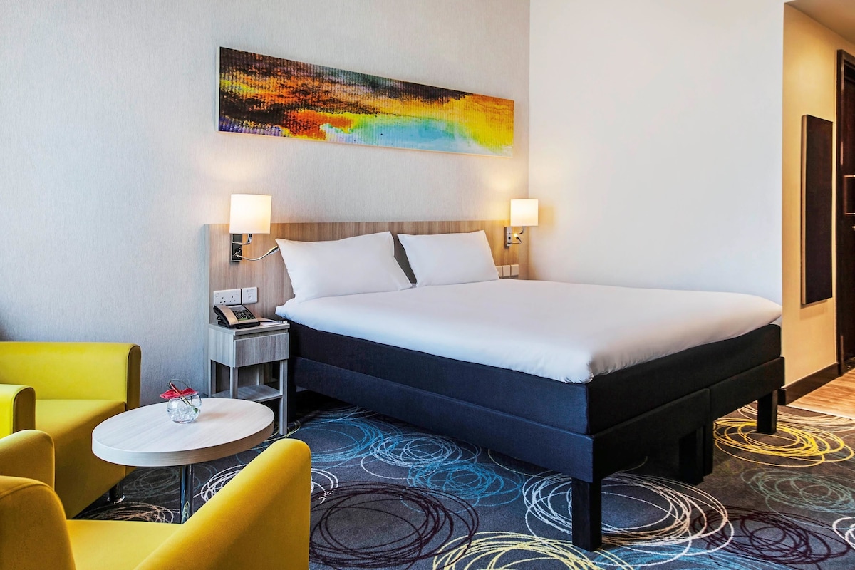 Een hotelkamer met een bed, nachtkastjes, een lamp, een stoel en een kleurrijk abstract schilderij aan de muur.