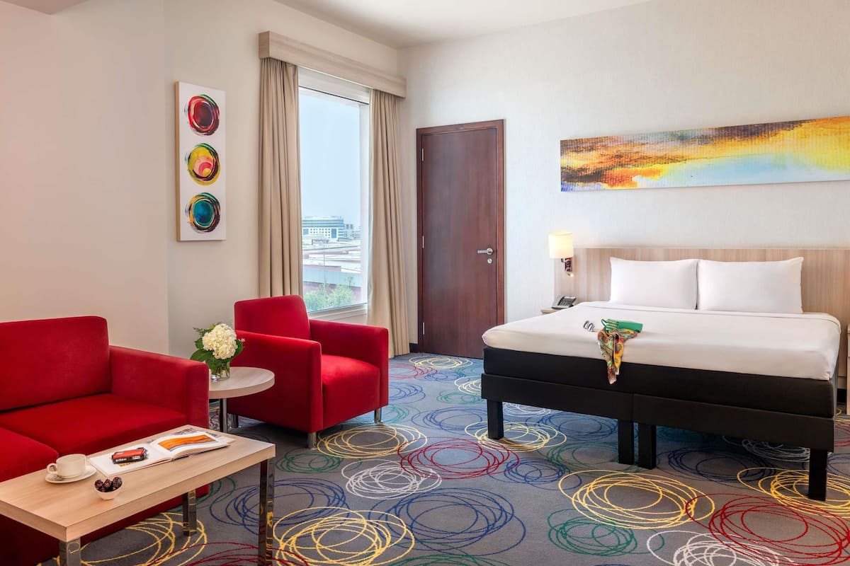 Une chambre d’hôtel avec un lit, un fauteuil rouge, une petite table avec un livre et une tasse, une moquette colorée et un tableau au mur.