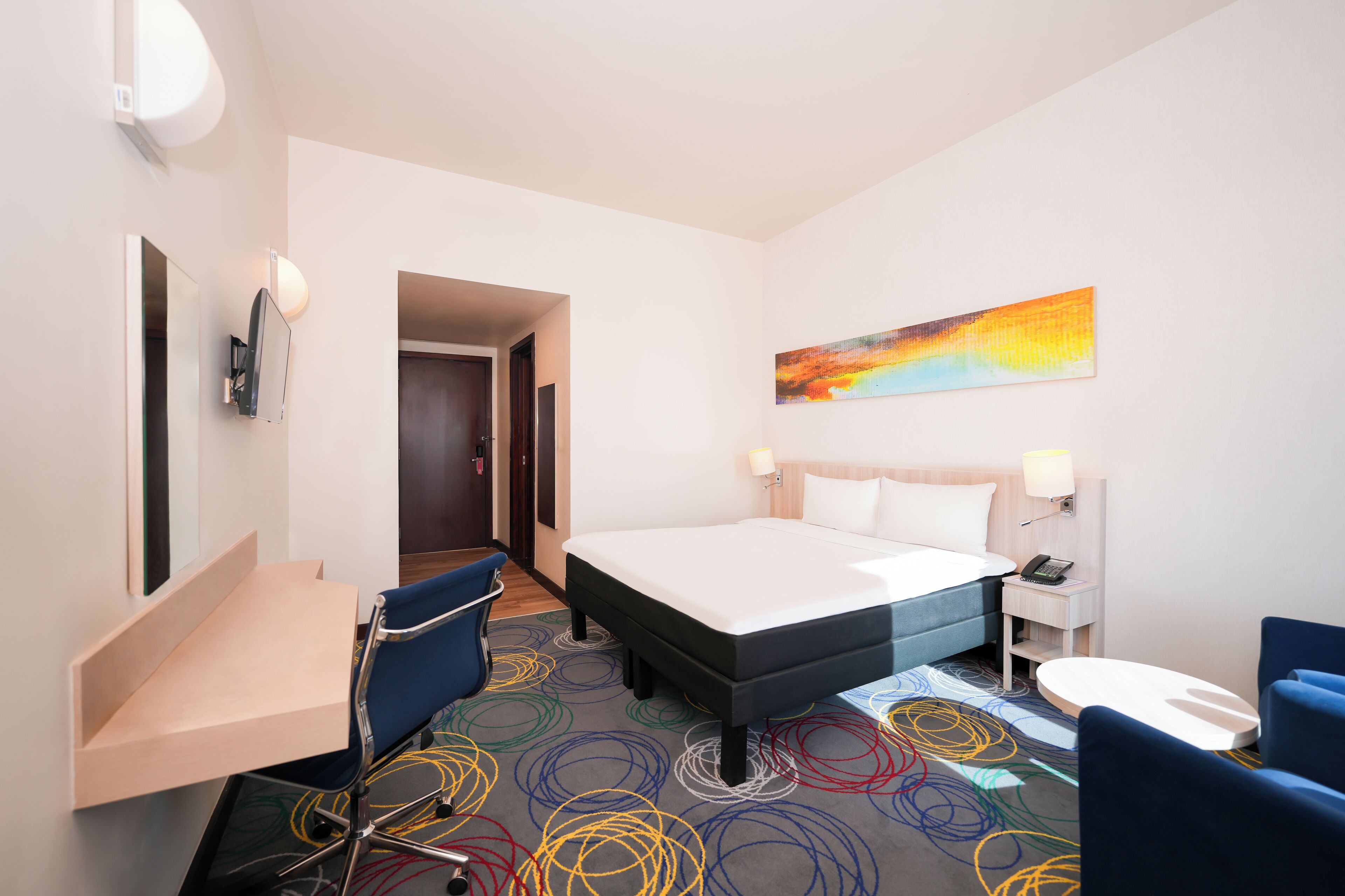 A hotel room with a bed, a desk, a chair, a small table, and a painting on the wall.