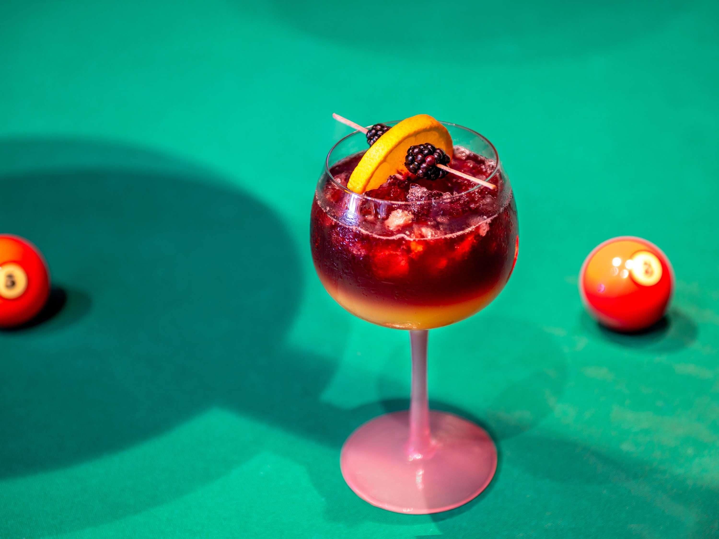 A cocktail in a glass with a fruit garnish, set against a green background with pool balls.