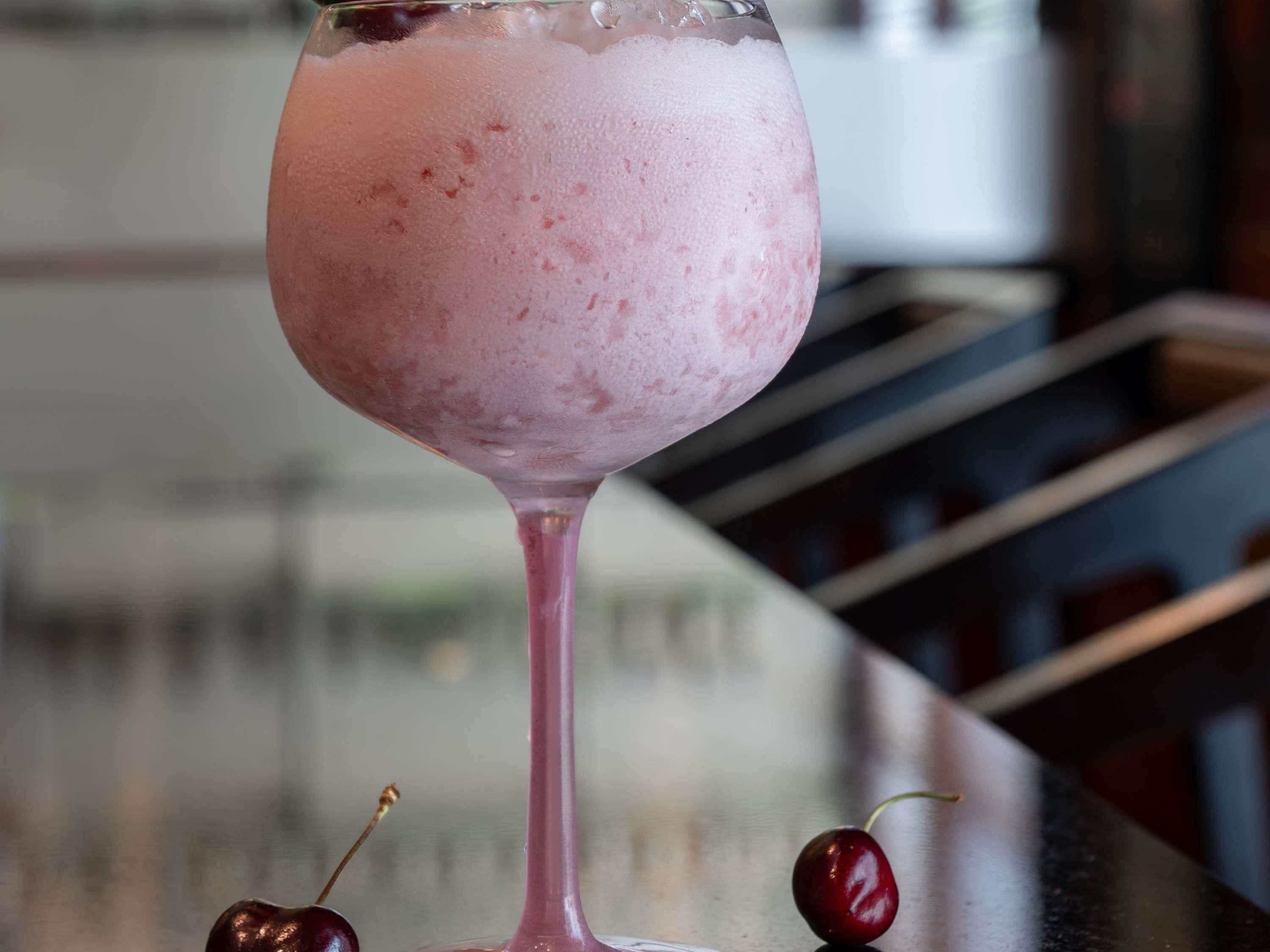 A pink drink in a glass with cherries on the bar counter.
