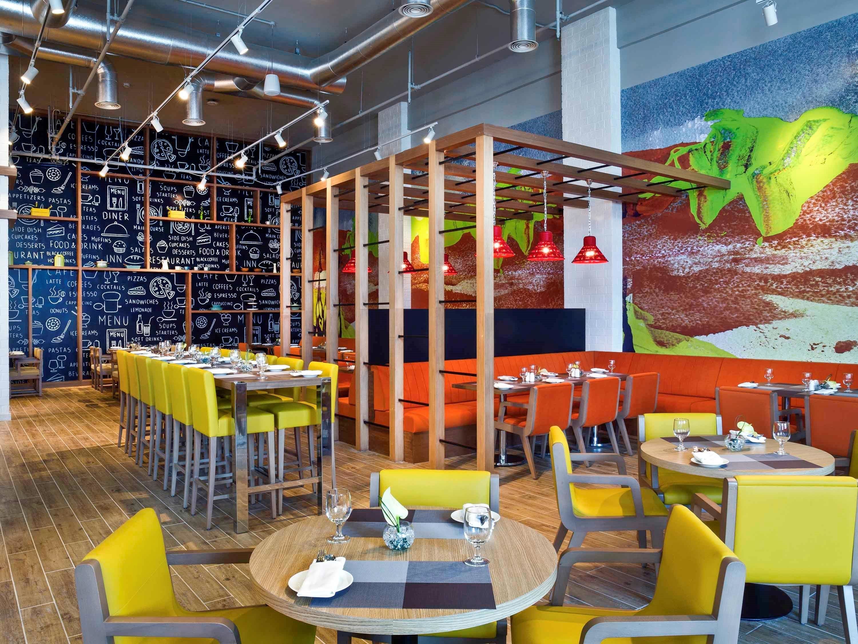Ein modernes Restaurant mit hohen Decken, freiliegenden Lüftungsrohren und einem lebendigen Wandgemälde. Tische sind mit gelben Stühlen und Holztischen gedeckt, und die Wände zeigen Kreidetafeln mit Speisen und Getränken.