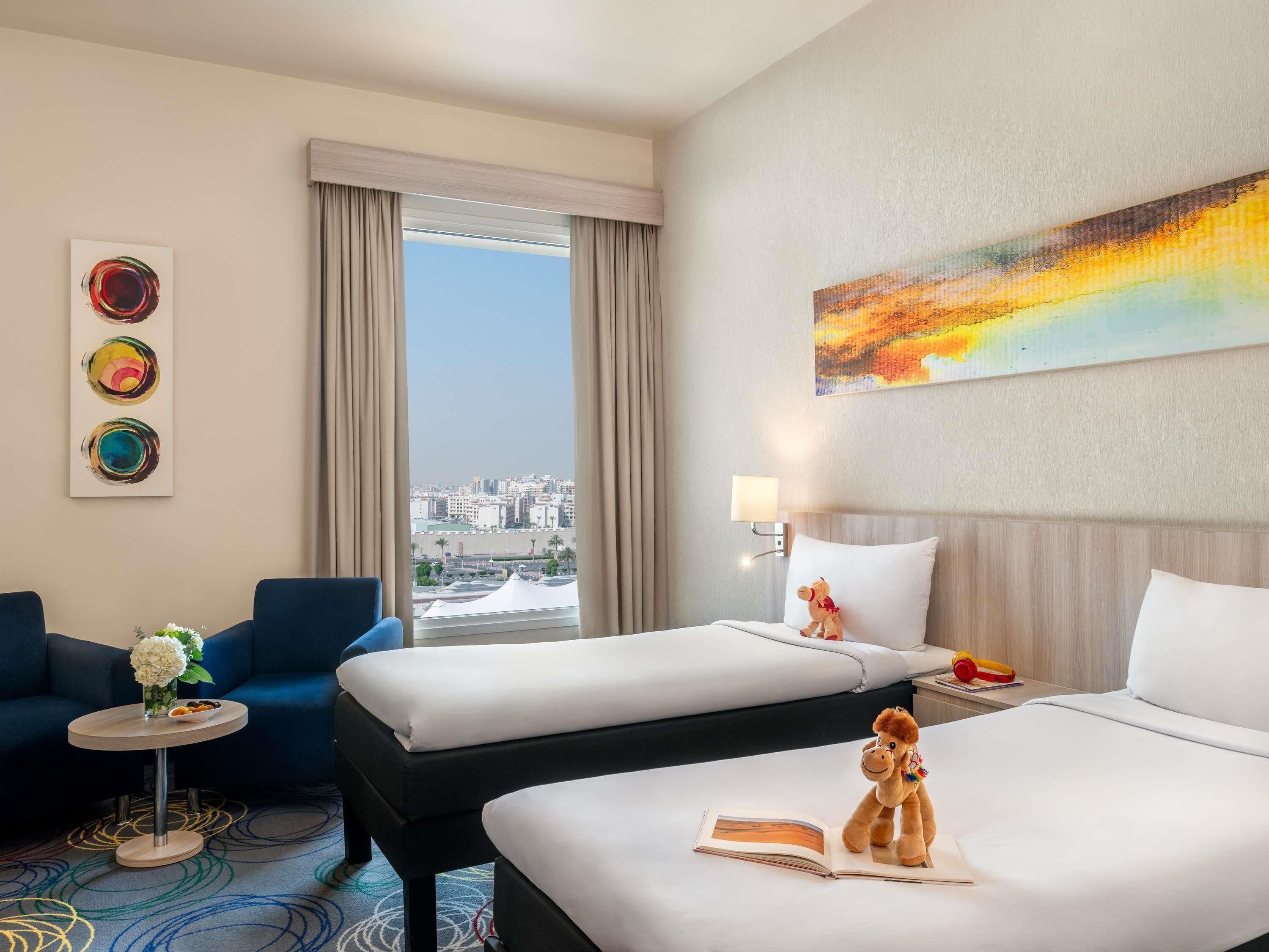 Ein Hotelzimmer mit zwei Betten, einem Schlafsofa, einem kleinen Tisch mit Blumen, einer Lampe und Blick auf die Stadt.