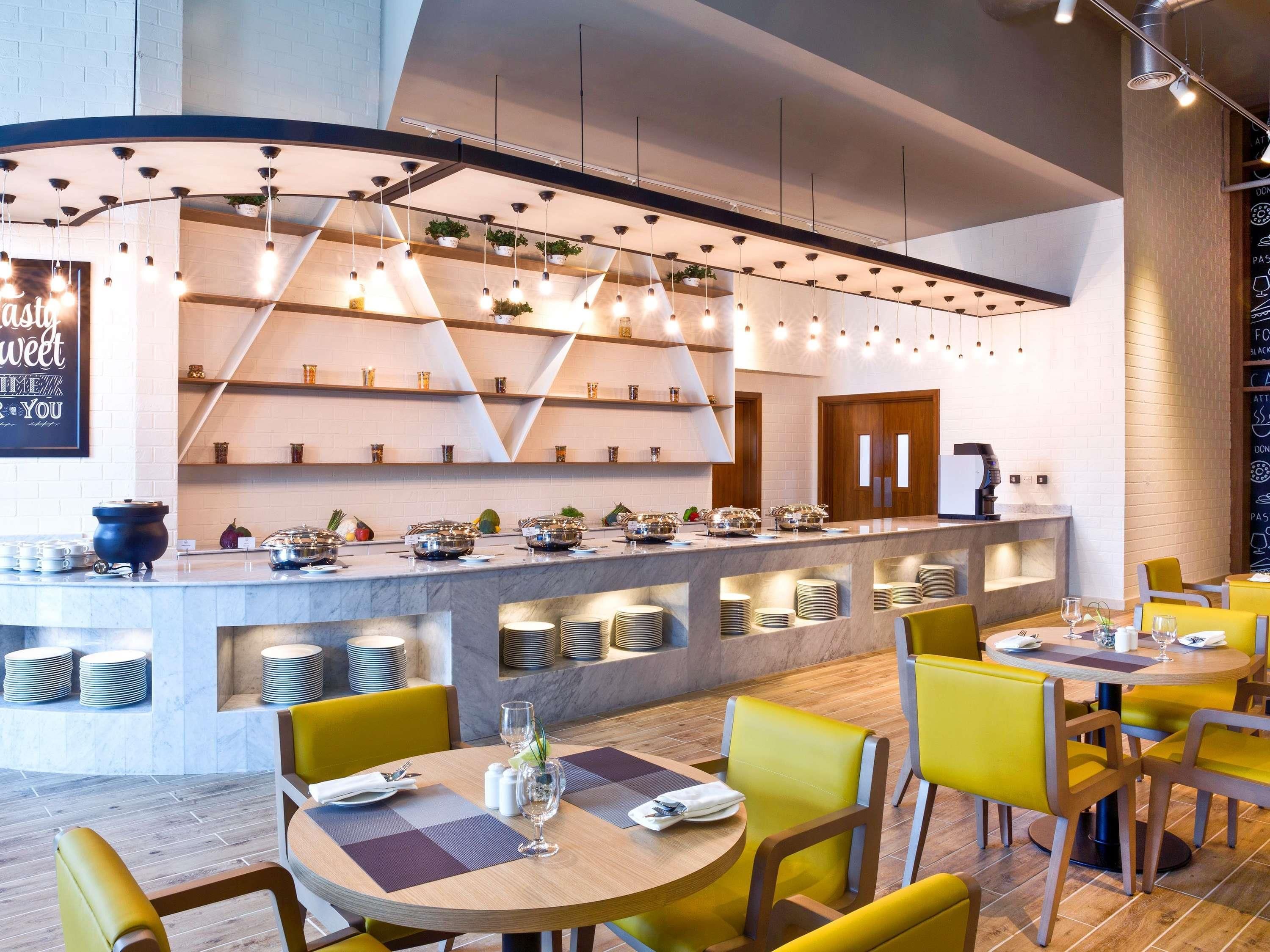 A modern restaurant with a central bar, yellow chairs, and a variety of dishes displayed on shelves.