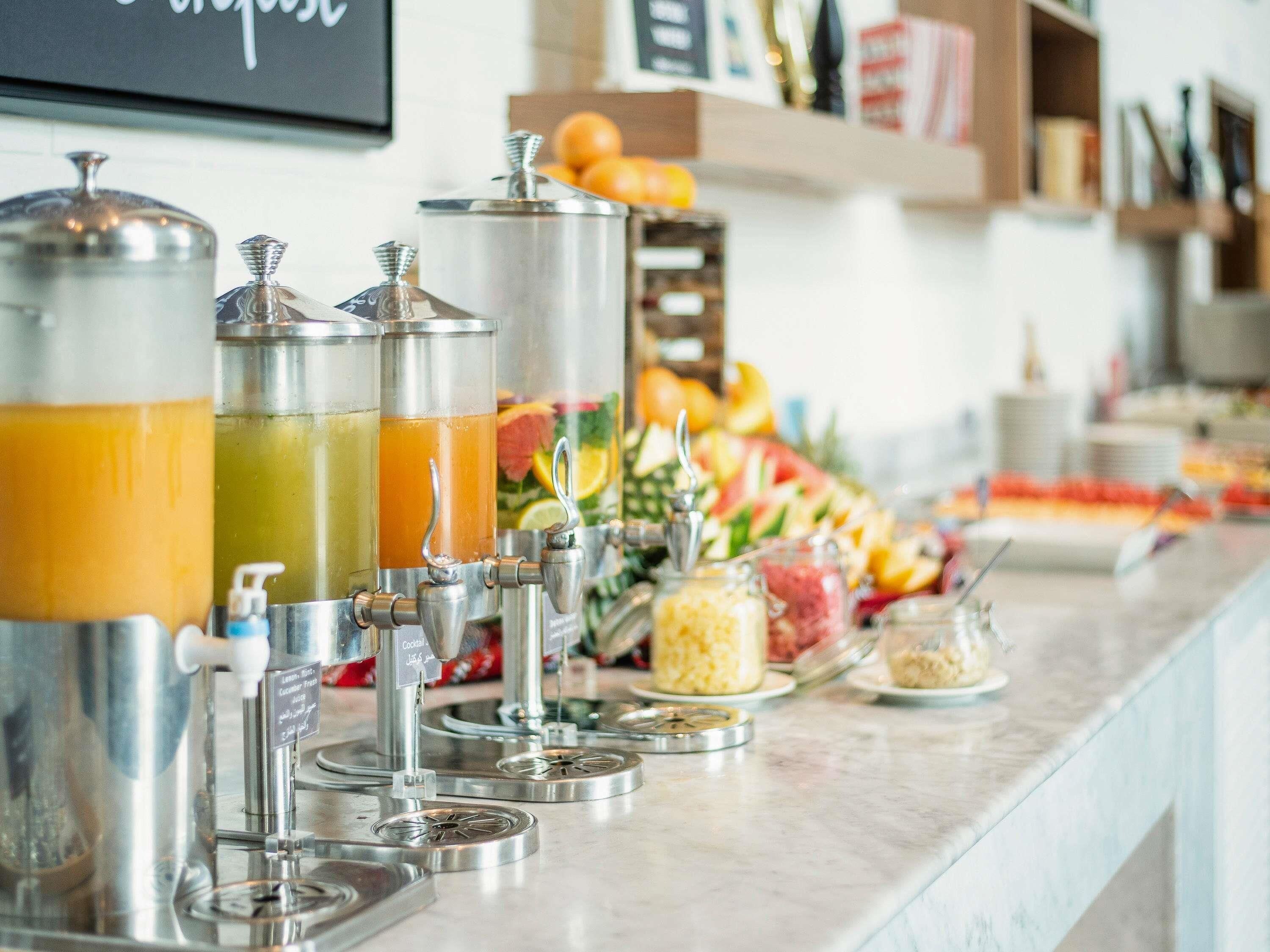 A breakfast bar with multiple beverage dispensers, a fruit display, and a variety of food items.