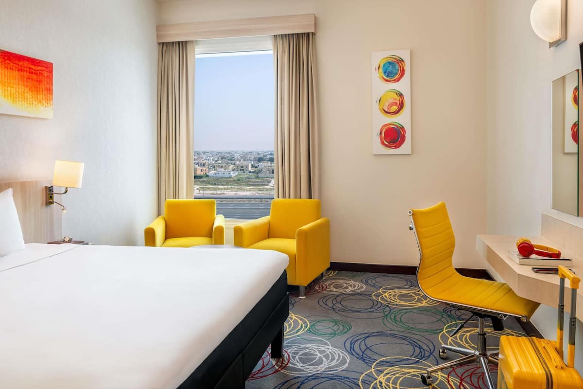 Une chambre d’hôtel comprenant un lit, deux fauteuils jaunes, un bureau avec une chaise, une fenêtre donnant sur un paysage urbain et des œuvres d’art abstraites accrochées au mur.