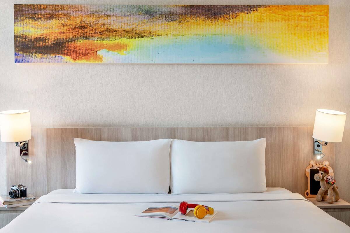 Een hotelkamer met een groot abstract schilderij, een bed met witte lakens, twee bedlampjes, een pluchen speeltje en een camera op het nachtkastje.