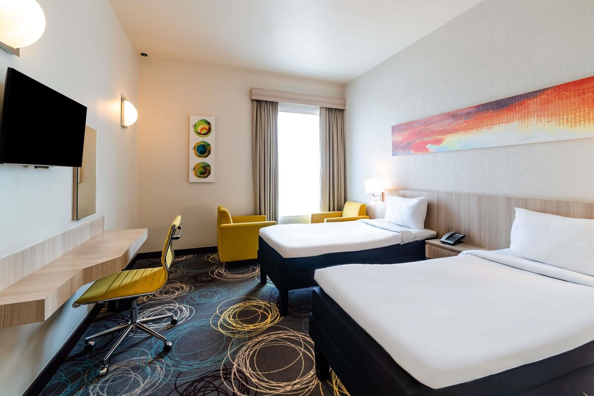 Une chambre d’hôtel avec deux lits, une télévision, un bureau avec une chaise et une fenêtre avec des rideaux.