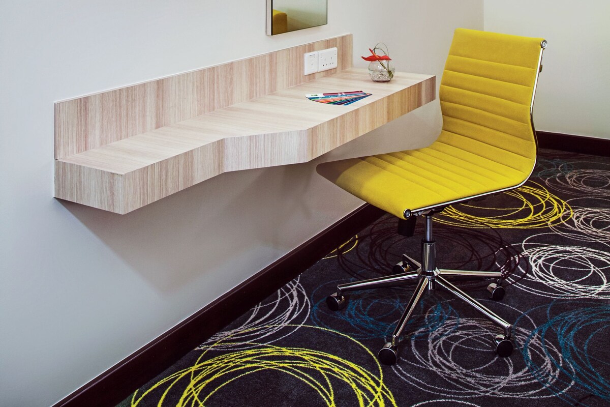 Een moderne hotelkamer met een wandtelevisie, een houten console, een gele stoel en een kamerbreed tapijt met een patroon.