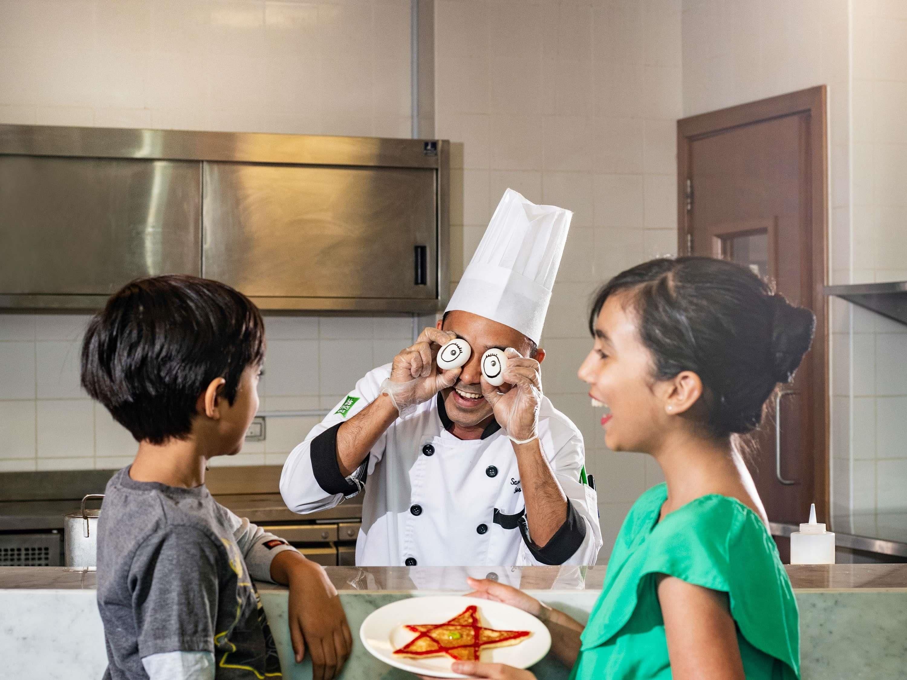 A chef in a white uniform with a tall hat, interacting with two children in a kitchen setting.