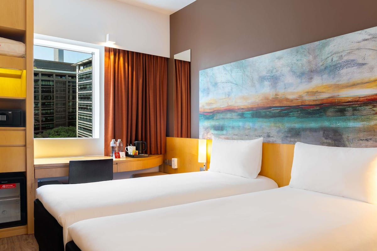 A hotel room with two beds, a desk with a computer, a window with curtains, and a large abstract painting on the wall.