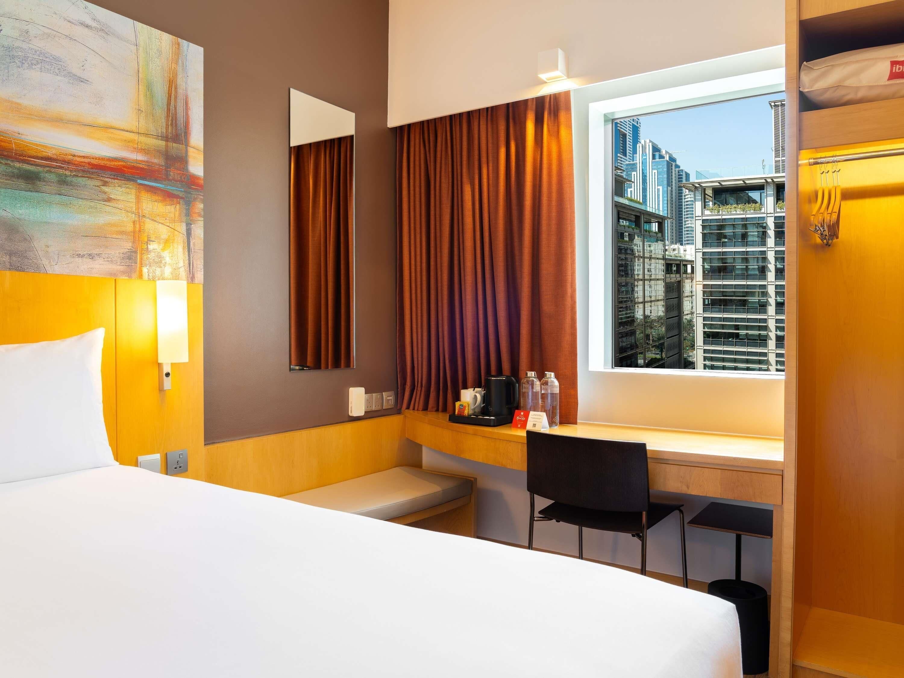 A hotel room with a bed, a desk, a chair, a window with a city view, and a painting on the wall.