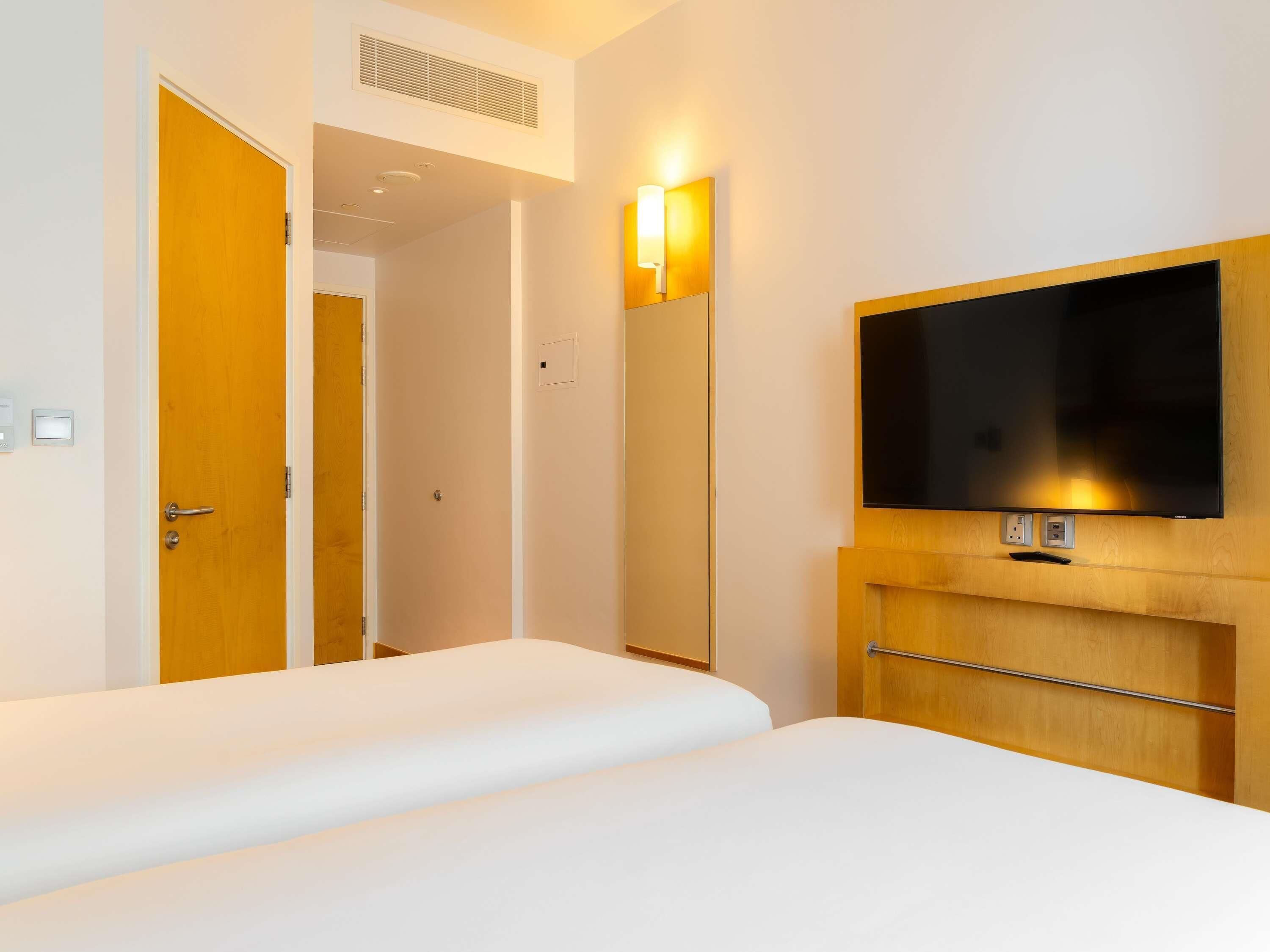 A hotel room with a bed, a TV mounted on a wooden panel, a door, and a light fixture.