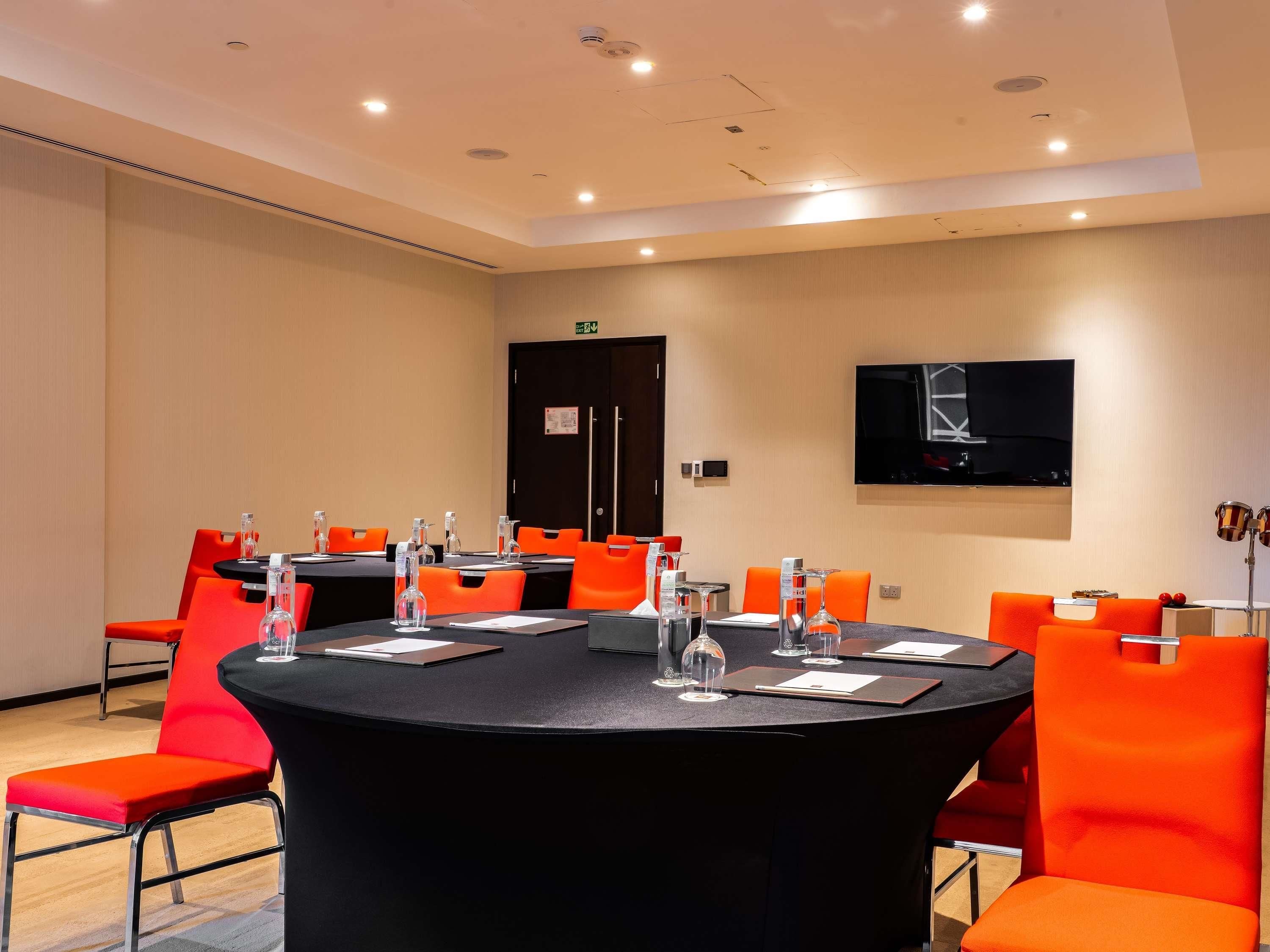 A conference room with a round table, orange chairs, a black tablecloth, and a mounted TV.