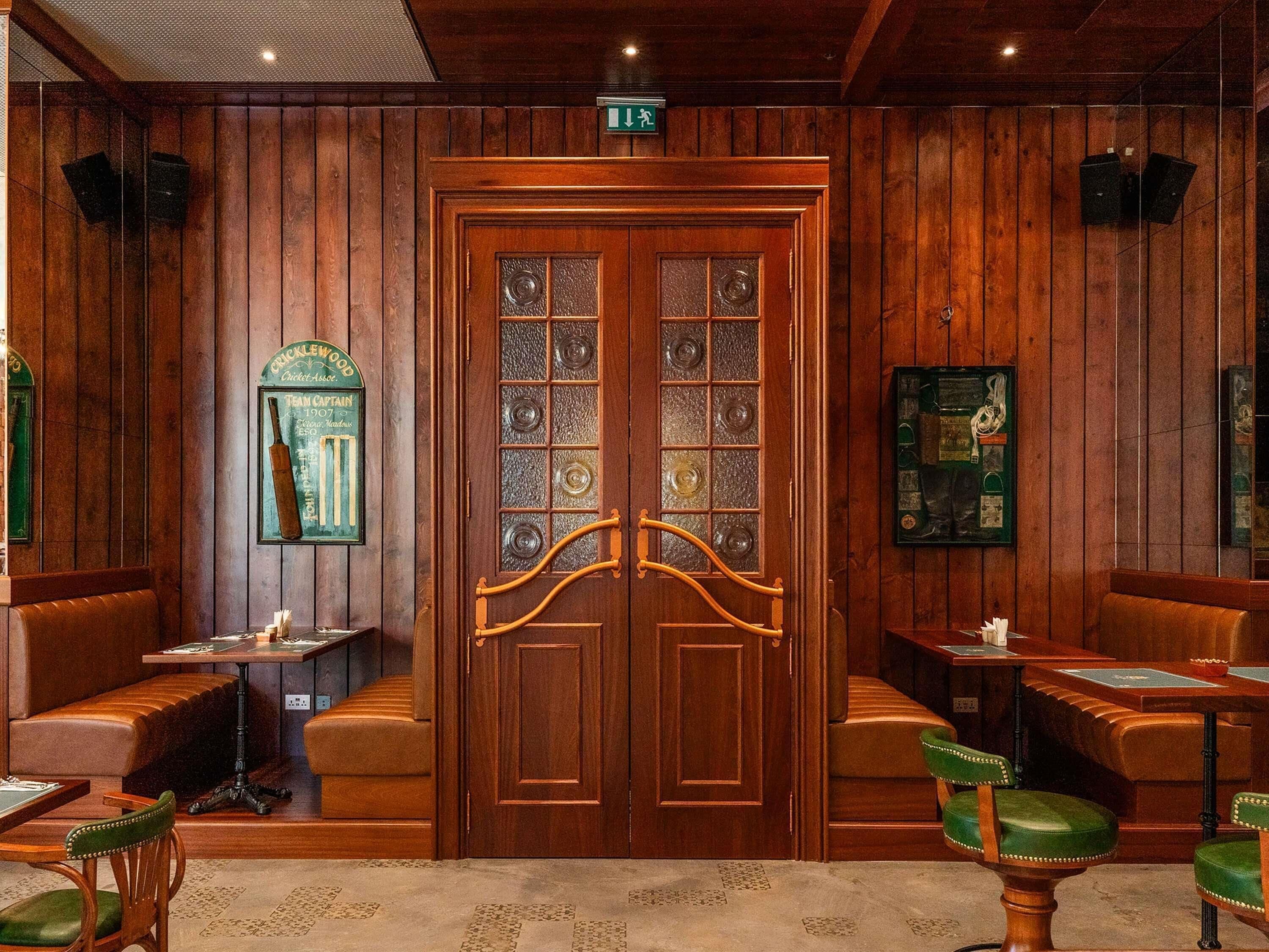 A wooden-paneled room with a large wooden door, two small tables with chairs, and a bench.