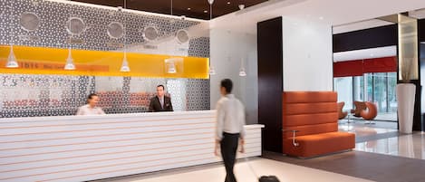 A modern hotel reception area with a patterned wall, a reception desk, and a seating area with orange chairs.