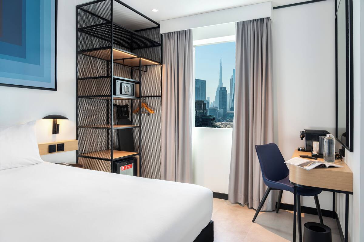 Une chambre d’hôtel moderne équipée d’un lit, d’un bureau, d’une chaise, d’une bibliothèque et offrant une vue sur le paysage urbain.