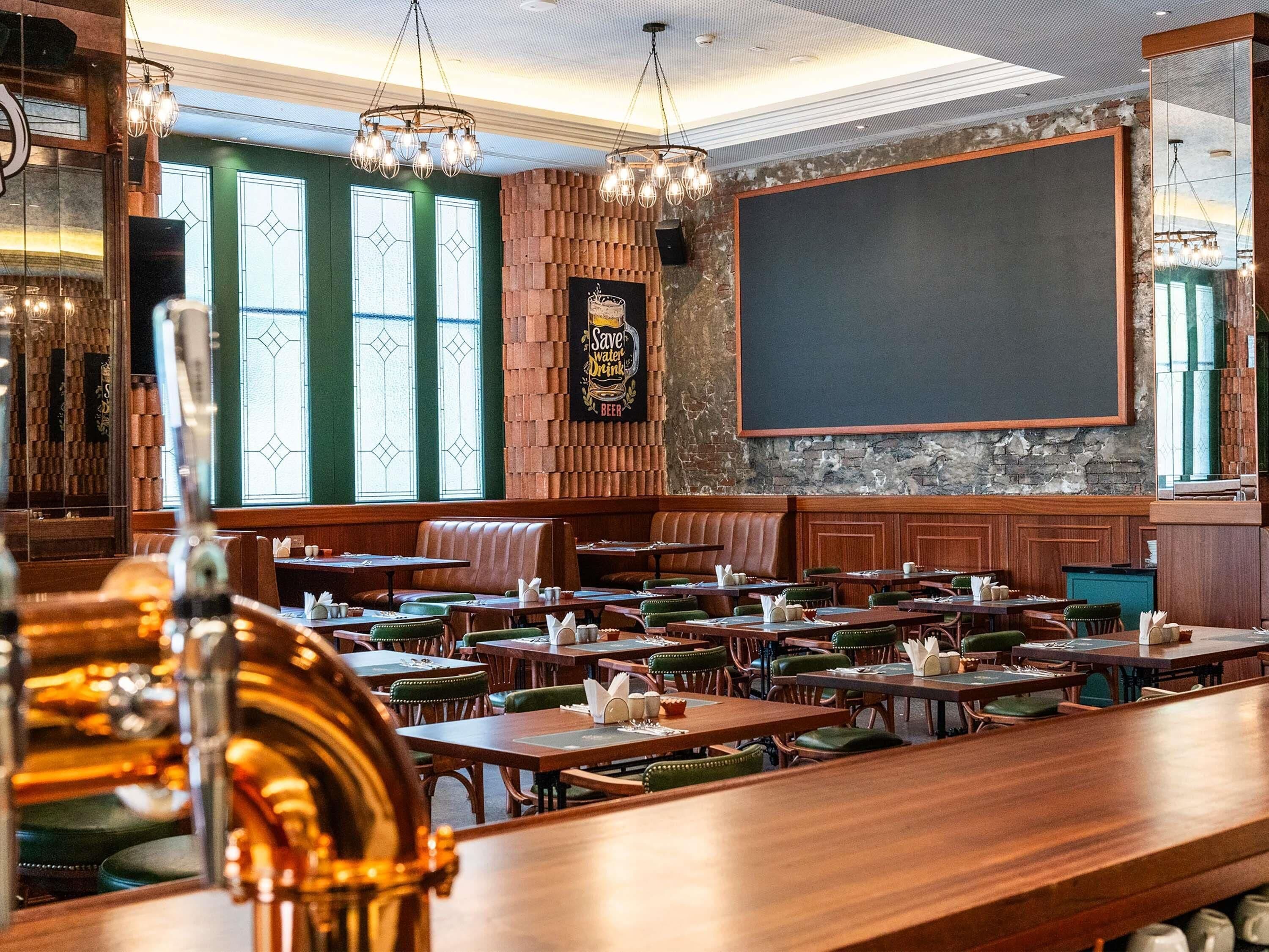 A restaurant with wooden tables and green upholstered chairs, a chalkboard wall, and a brick wall with a 'Save Water Drink Beer' sign.