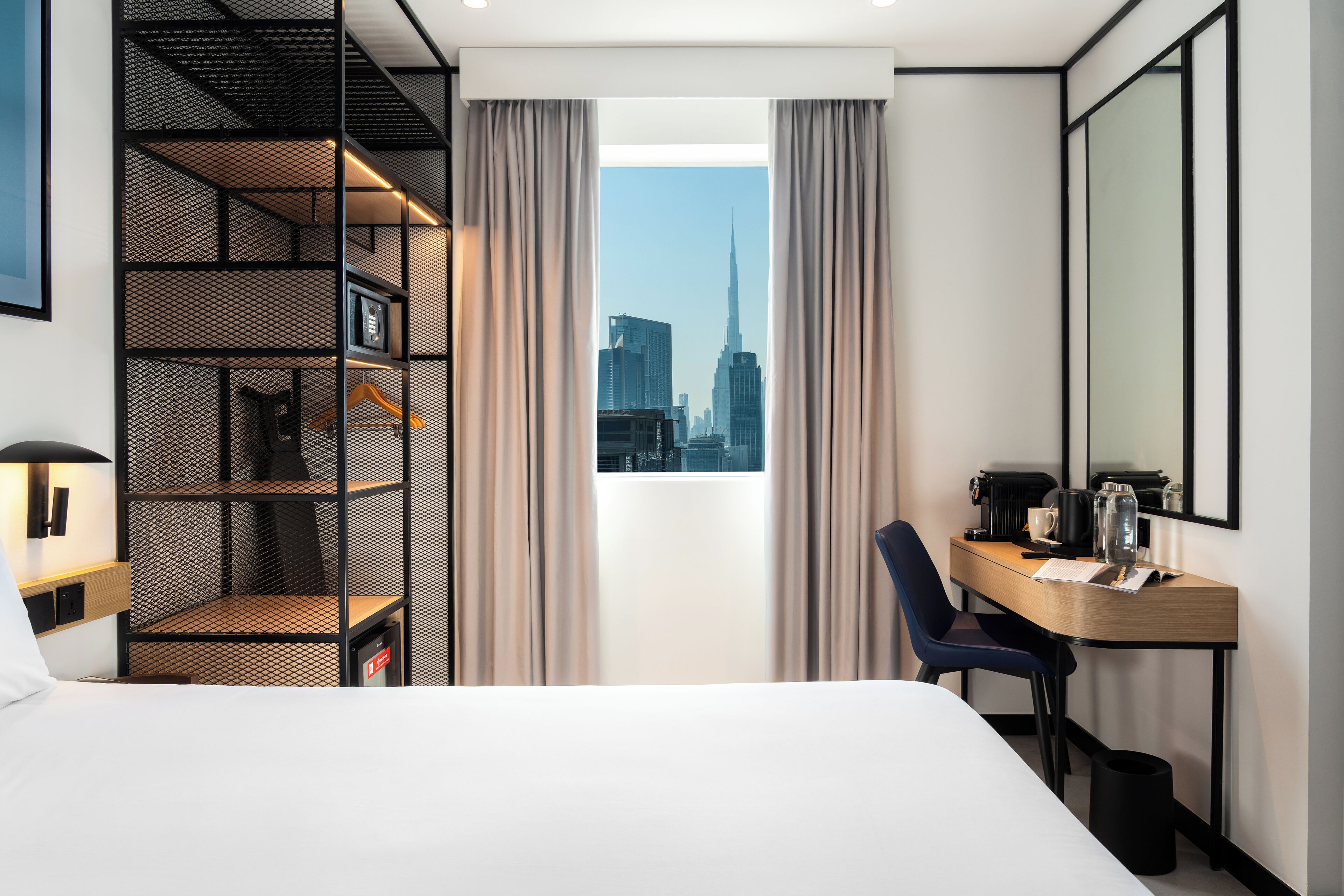 A hotel room with a bed, a desk with a chair, a bookshelf, and a large window with a city view.