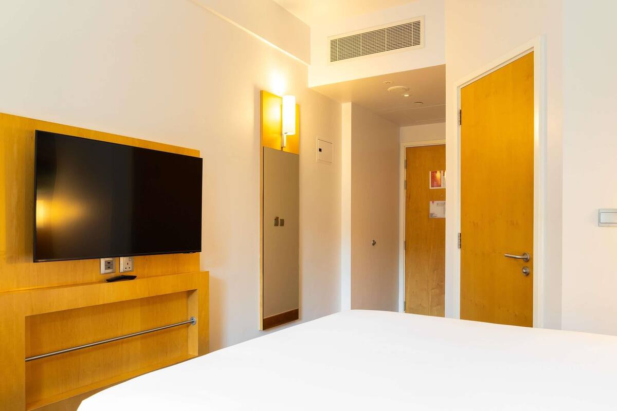 Hotellihuone, jossa on suuri sänky, seinälle asennettu televisio, puinen kaappi ja ovi, joka johtaa toiseen huoneeseen.