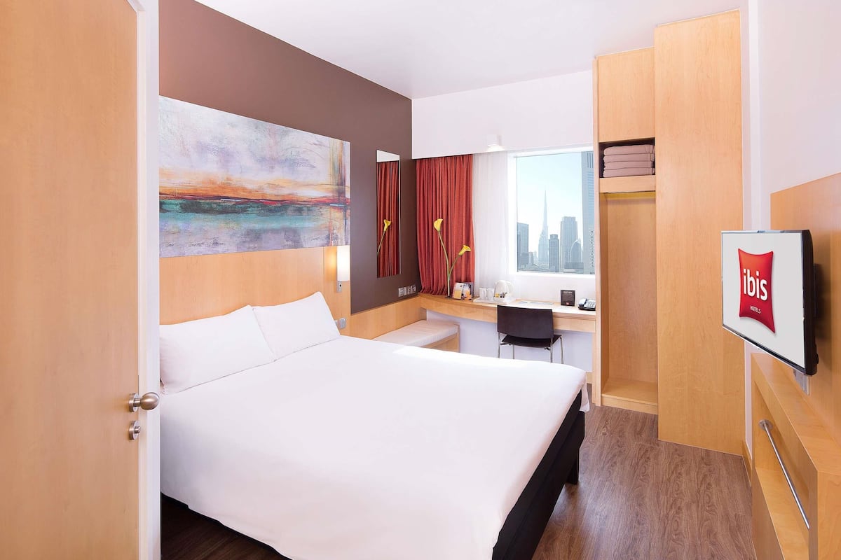 A hotel room with a bed, desk, chair, artwork, window, and a TV displaying the ibis logo.
