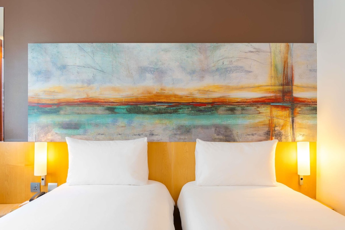 Een hotelkamer met twee bedden, een groot abstract schilderij aan de muur en twee bedlampen.