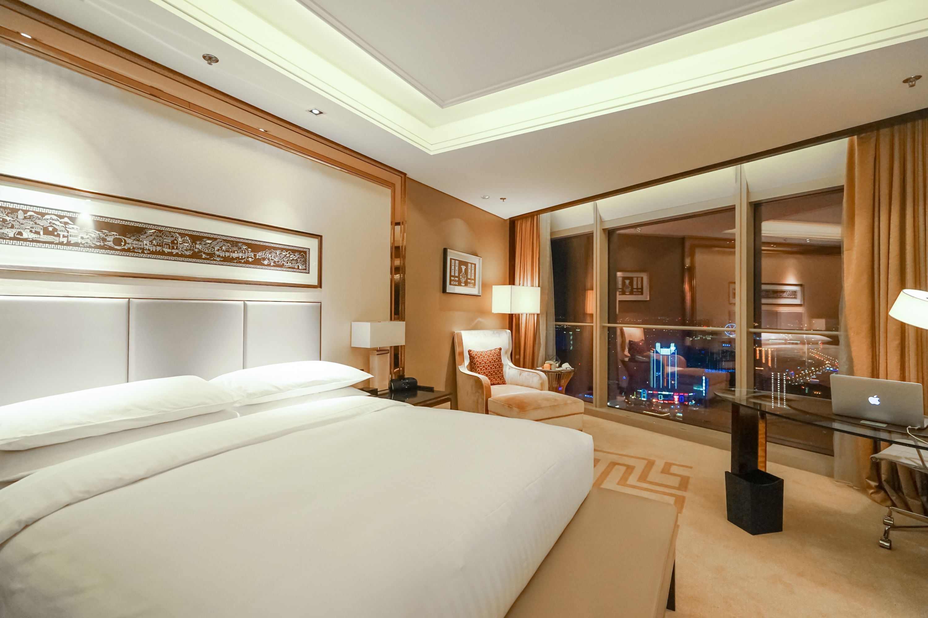 Habitación de hotel con una cama grande, un escritorio con una computadora portátil, una silla y vistas de la ciudad a través de un ventanal amplio.