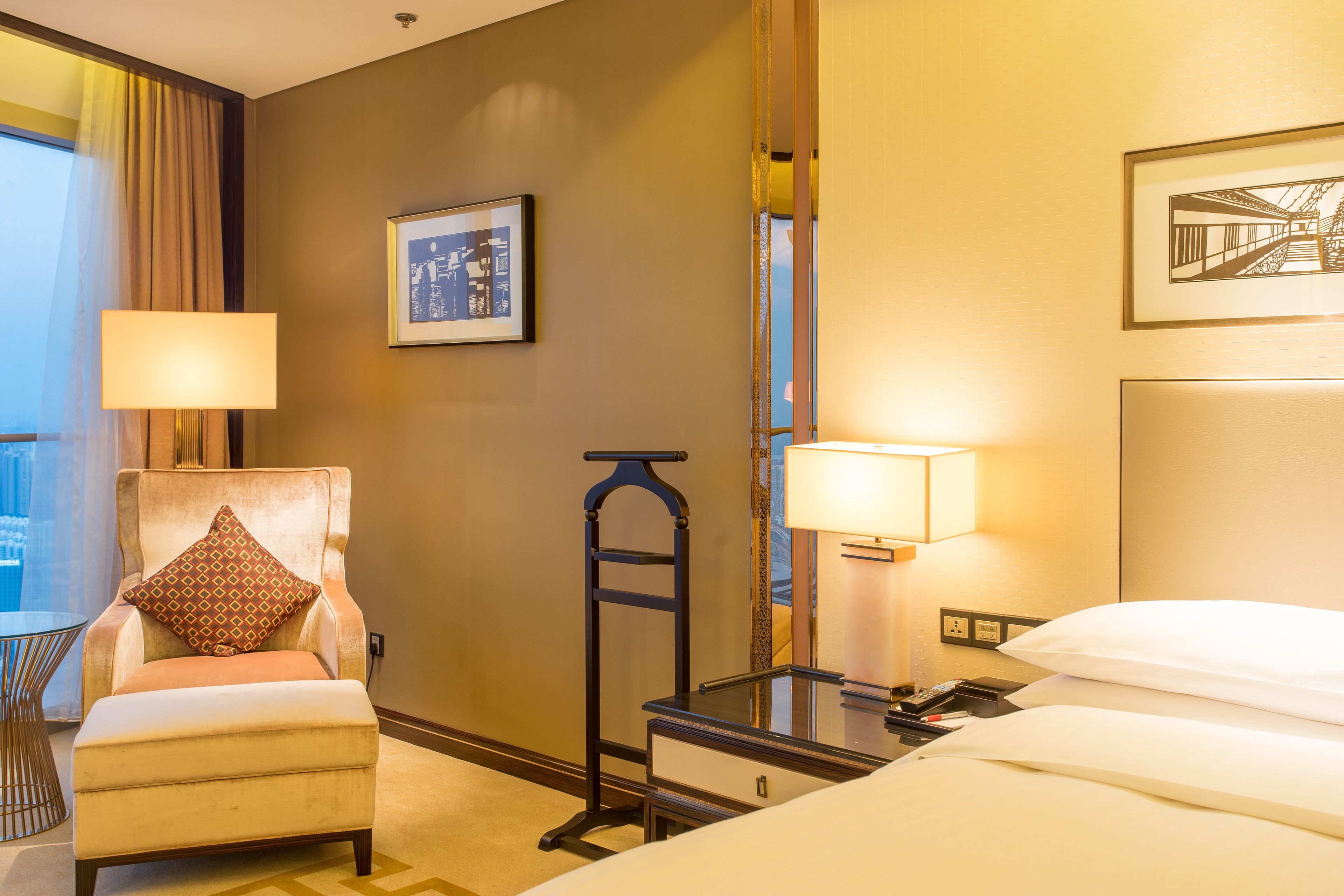A hotel room with a bed, a chair with a pillow, a bedside table with a lamp, a mirror, and a framed picture on the wall.