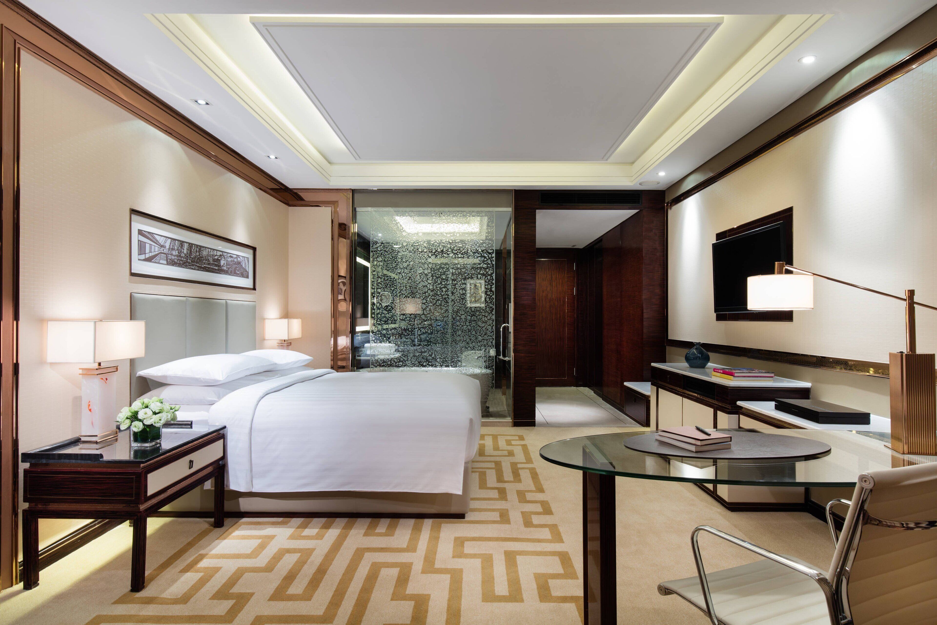 A modern hotel room with a large bed, bedside tables, a glass coffee table, a chair, a TV, and a bathroom with a glass shower.