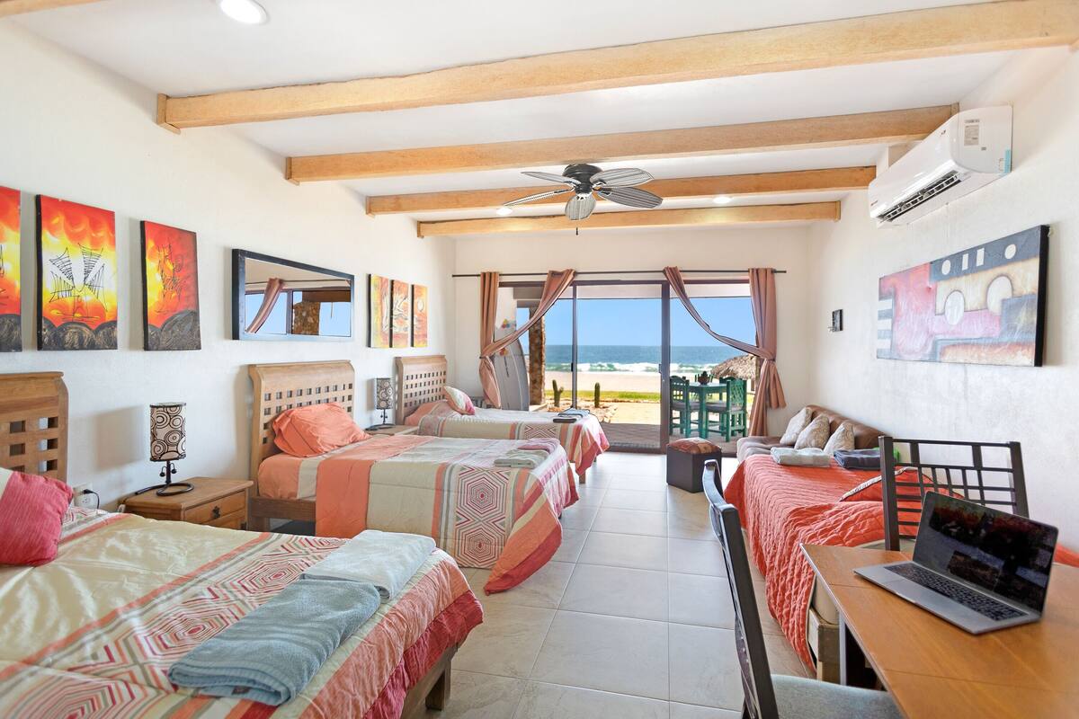 A hotel room with three beds, a laptop on a desk, and a view of the beach.