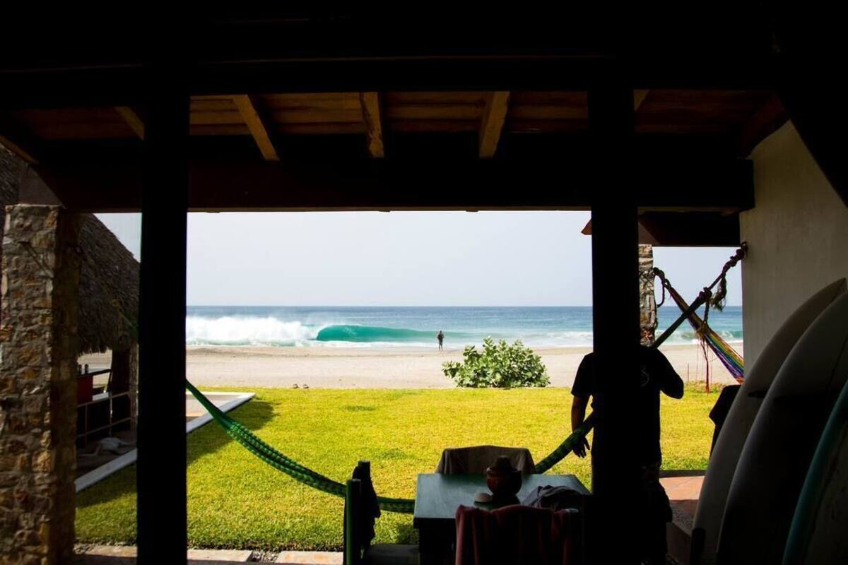 A beach view from a covered area with a table and chairs, a surfboard, and a rope.