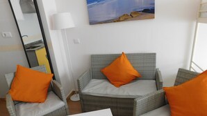 A room with wicker furniture, including two armchairs and a sofa, each with orange cushions. There is a white table in the center. A painting of a beach scene is on the wall, and a floor lamp is visible. - Cádiz INN Hostel (Cádiz)